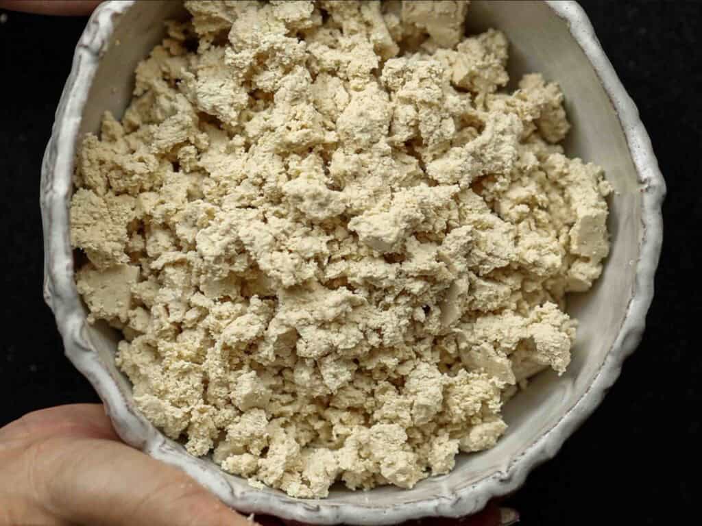 A hand holds a ceramic bowl filled with crumbled tofu, perfect for vegan bolognese, against a dark background. The tofu pieces vary in size and have a light beige color.